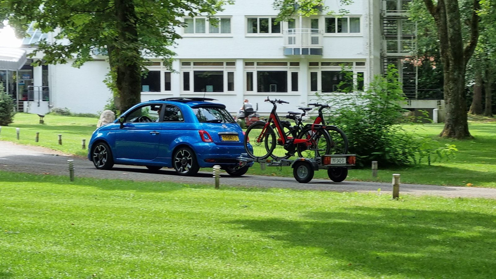 Blauwe auto met een trailer waarop twee rode fietsen staan, in een park